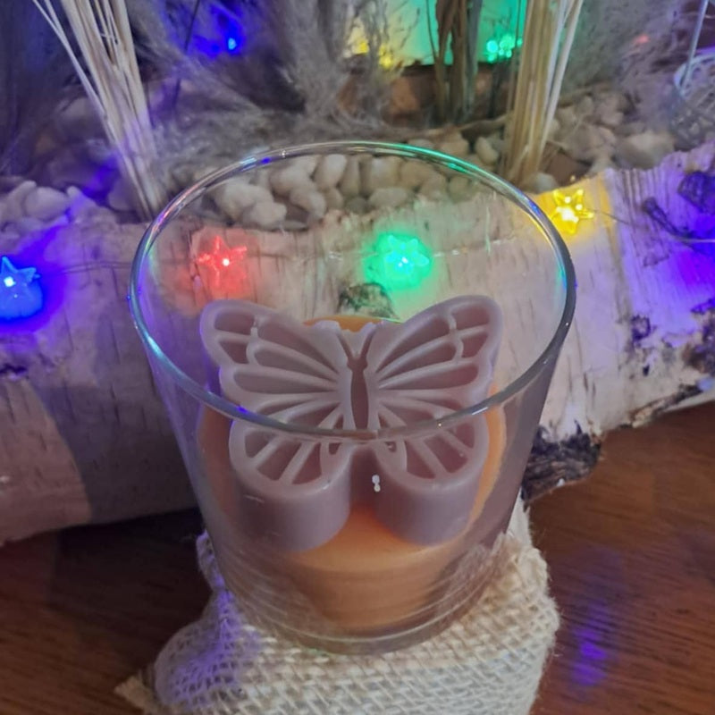 Bougie artisanale en verre avec un papillon rose sculpté sur le dessus, nommée L'Éveil d'Elya, symbole de renouveau et d'espoir.