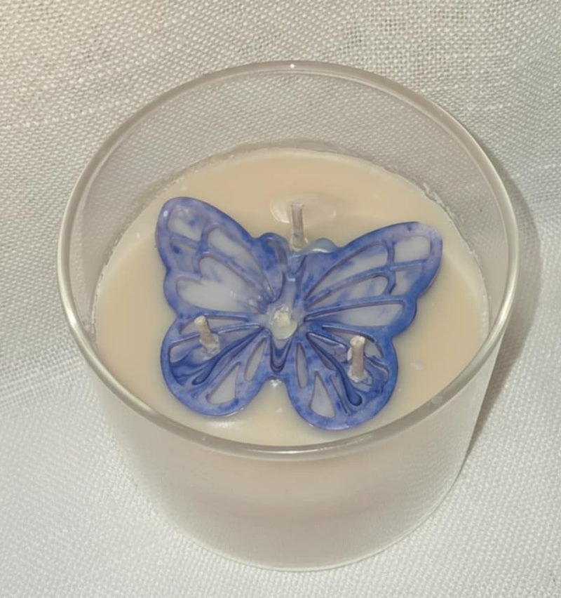 Bougie artisanale en verre avec un papillon violet sculpté sur le dessus, nommée L'Éveil d'Elya, symbole de renouveau et d'espoir.